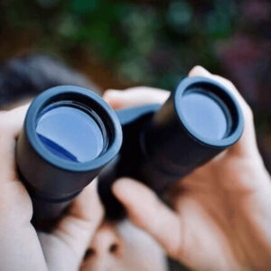 Närbild av en person som tittar i en kikare. Endast händerna, kikaren, näsan och ena kinden syns, tillsammans med suddigt mörkt hår och bakgrund. Fokus ligger på kikarens glas som blänker. Bilden används som produktbild till jobbpaket-plus för arbetsgivare.