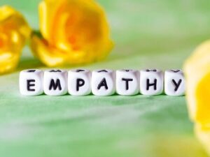 Sju tärningar på rad med bokstäver som bildar ordet "Empathy". I bakgrunden syns två gula rosor mot en ljusgrön, suddig bakgrund. Bilden symboliserar ett av Socialstyrelsens kompetensmål för undersköterskor.