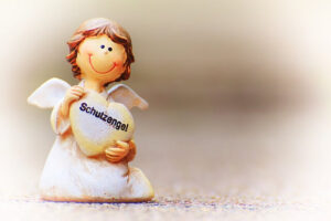 En liten glad skyddsängel av porslin med brunt hår, som ligger på knä och håller i ett hjärta där det står "Schutzengel" på tyska. Bakgrunden är suddig. Bilden används för att uppmuntra till att bli god man, förvaltare eller särskilt förordnad vårdnadshavare.