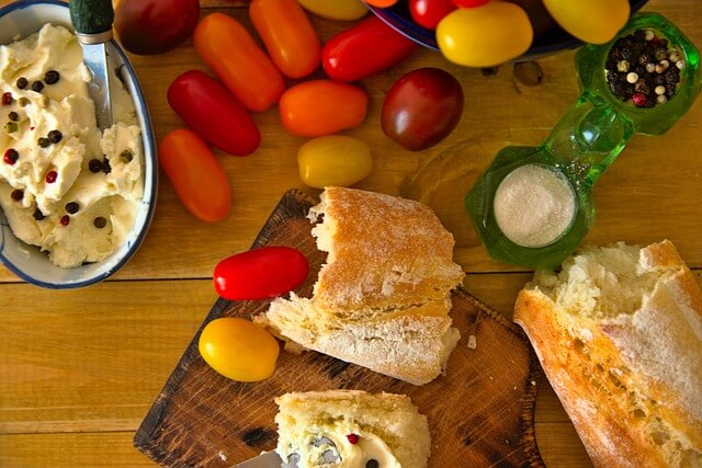 Baguette bruten i bitar på en skärbräda. Bakom brödet, på träbordet, syns färgglada tomater och olika skålar med innehåll. Bilden bidrar till sidans visuella design.