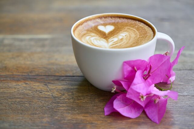 Vit kaffekopp på ett brunt träbord. Koppen är fylld med kaffe och täcks av cappuccinoskum, där ett hjärta och vågformationer har skapats. Bredvid koppen ligger en lila bougainvillea-blomma. Bilden bidrar till namnsdags-inläggets visuella design.