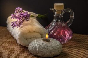Brunt underlag med tydlig, lite grövre textur. En ihoprullad vit handduk till vänster i bild med en kvist av blålila lavendelblommor liggande på handduken. Till höger en vacker glaskaraff med lilarosa innehåll. I förgrunden en stenljusstake med en brinnande låga. Bilden bidrar till namnsdags-inläggets visuella design.