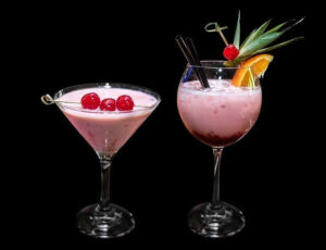 Två cocktailglas fyllda med rosa innehåll mot en mörk bakgrund. Till vänster ett trattformat glas garnerat med tre röda cocktailbär på en pinne, och till höger en kupa med svart sugrör, en cocktailpinne med blad, ett rött cocktailbär och en apelsinklyfta på glaskanten. Bilden bidrar till namnsdags-inläggets visuella design.