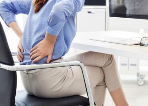 En kvinna sitter på en stol och håller händerna över ländryggen som tecken på ryggsmärta. Hon är klädd i ljusblå skjorta och beige byxor och sitter med benen i kors på en stol med kromat stål. Bakgrunden visar en vit skrivbordsskiva och en del av en datorskärm. Bilden används i ett inlägg om risken för belastningsskador.