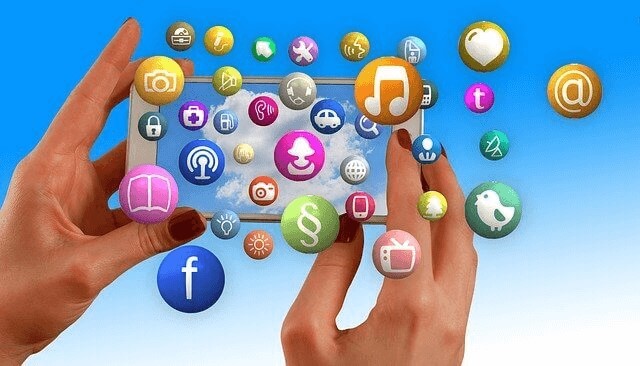 Två händer håller en silverfärgad mobil som visar en ljusblå skärm med ett vitt moln. Färgade ikoner för sociala medier och verktyg strömmar ut från skärmen.