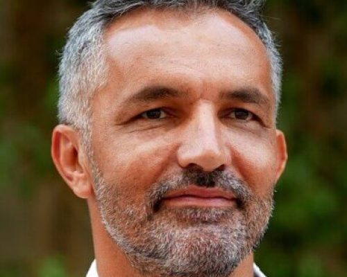 Porträtt av en man i yngre medelåldern med kortklippt grått hår och silverfärgade tinningar. Han har mörka ögon samt ett kort skägg och mustasch med grått och silverinslag. Han bär en vit skjorta och bakgrunden är suddig i grönbruna toner.