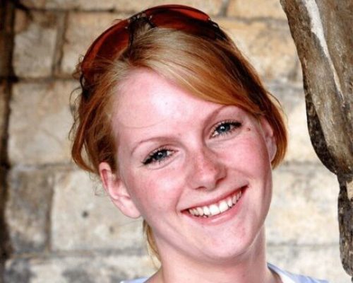 Porträtt av en ung kvinna med rödblont hår och snedlugg som ler stort så att tänderna syns. Hon har ljus hy och hennes hår är uppsatt i nacken. Hon bär en vit arbetsskjorta, typisk för vårdpersonal. Bakgrunden är en vägg i grå, gula och beige toner med tegelstensliknande block.