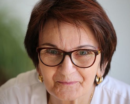 Porträtt av en medelålders kvinna med kort, mahognyfärgat hår och matchande glasögon. Hon bär guldringar i öronen och en vit arbetsrock, där enbart axlarna syns. Bakgrunden är suddig i grågröna toner.