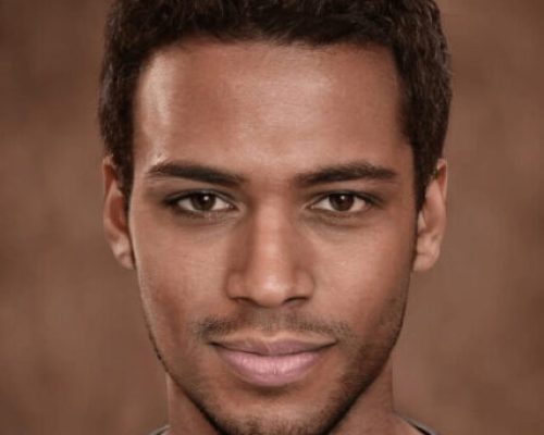 Porträtt av en yngre mörkhyad man med kortklippt mörkt hår, riktigt kort skägg och mustasch. Han har mörka ögon och bär en grågrön t-tröja, vars axlar skymtar. Bakgrunden är nästan i samma bruna ton som hans hy.